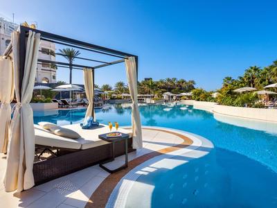 Großer blauer Pool mit Sonnenliegen, weißen Vorhängen und Palmen unter klarem Himmel.