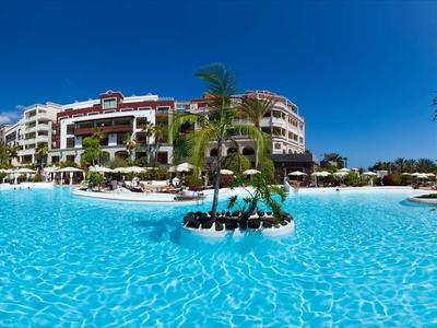 Velký bazén s deštníky a palmami před hotelovou budovou pod modrou oblohou