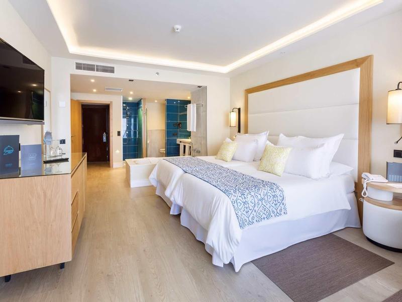 Camera d'hotel ben illuminata con due letti, TV, scrivania e bagno moderno.