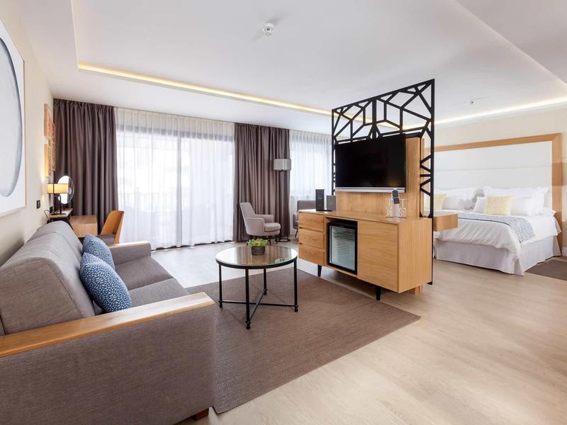 Camera d'hotel luminosa con area salotto, divisorio TV moderno e letto matrimoniale grande.