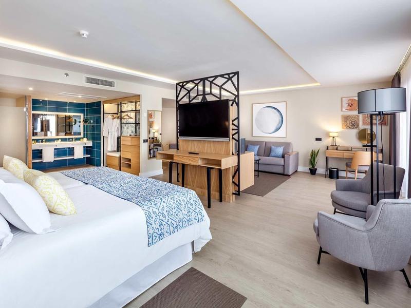 Camera d'hotel moderna con letto, scrivania, zona lounge e grande parete in vetro verso il bagno.