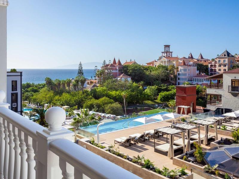 Vista da un balcone che domina una piscina, un giardino, edifici e il mare sullo sfondo.