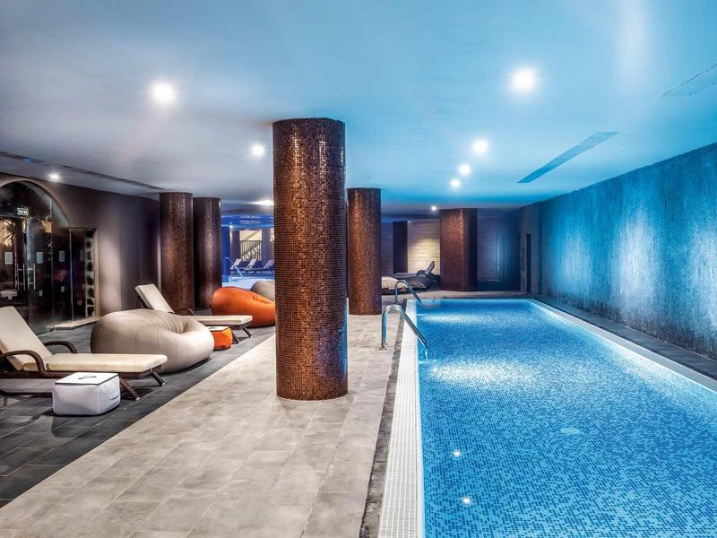 Piscina interna con diverse sedie a sdraio e illuminazione moderna in un hotel.