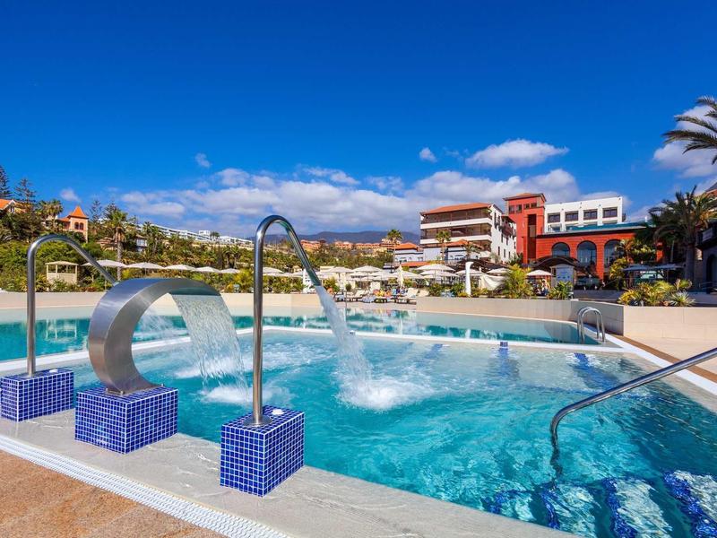 Piscina esterna moderna con getti d'acqua e edifici dell'hotel circostanti sotto un cielo blu.