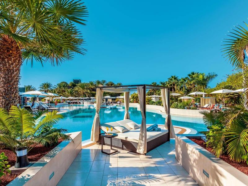 Area piscina di lusso con lettini eleganti, palme e cielo azzurro limpido.