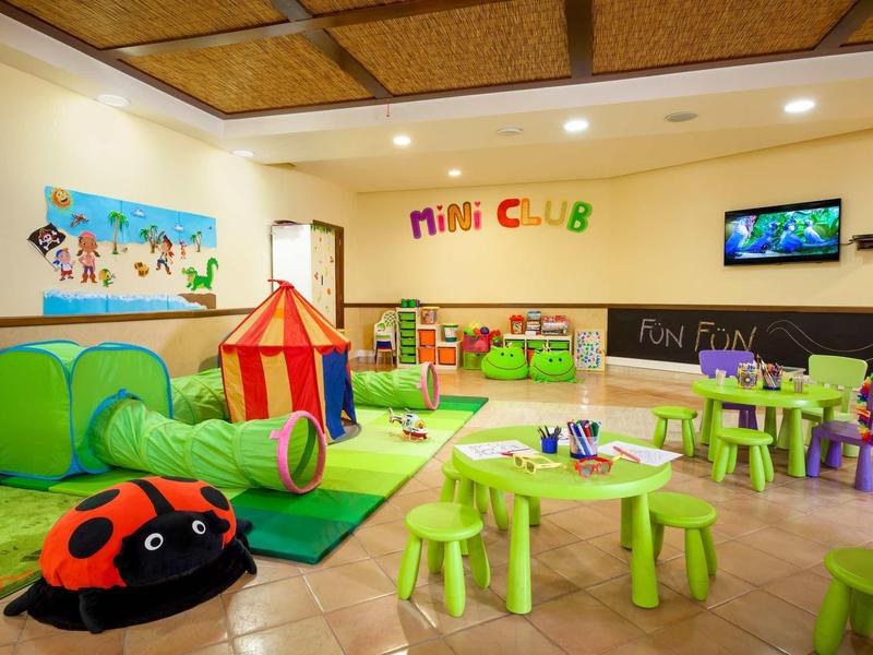 Sala giochi colorata per bambini con tende da gioco, piccoli tavoli e sedie, decorazioni murali e TV.