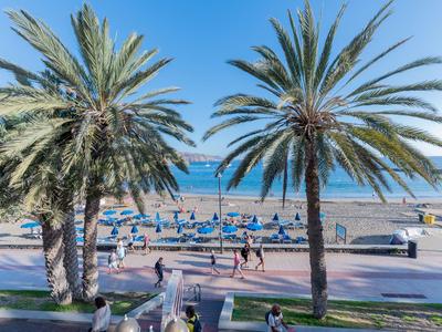 Vista de la playa con palmeras, sombrillas azules y gente paseando por un paseo marítimo.