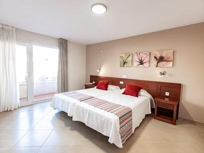 Habitación de hotel luminosa con dos camas individuales, mesitas y acceso al balcón por ventanas grandes.