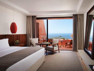 Modernes Hotelzimmer mit großem Bett, Sessel, Tisch und Balkon mit Meerblick und Sitzmöbeln.