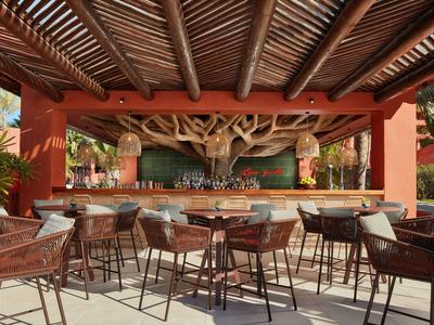 Offene Bar mit Holzstühlen, Tischen und großer Baumdeko unter einem Holzdach im Freien.