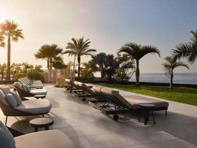 Atardecer sobre una terraza con piscina con tumbonas y palmeras junto al mar.