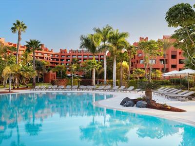 Großer Pool mit Sonnenschirmen und Palmen vor einem roten Hotelgebäude bei blauem Himmel.
