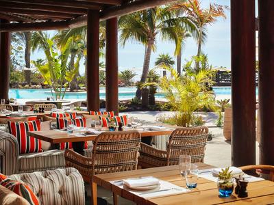 Zona exterior de restaurante con muebles de madera con vista a la piscina y palmeras.