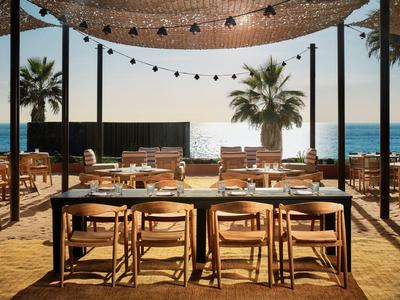 Strandrestaurant mit Holztischen, Stühlen, Palmen und Meer im Hintergrund bei Sonnenuntergang.