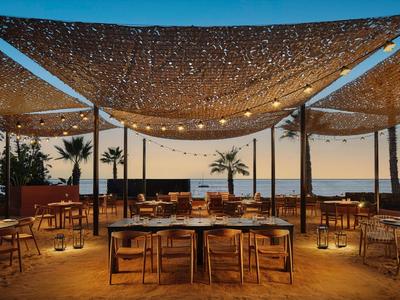 Cena al aire libre bajo toldos beige con vista al mar y palmeras a lo lejos.