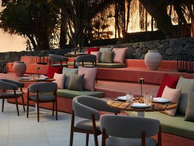 Gemütliche Außenlounge mit roten und grauen Polstern, Tischen und Dekoration in warmem Licht.