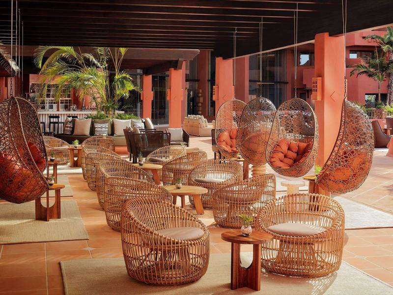 Moderne Lounge mit geflochtenen Sitzmöbeln, Hängekörben und warmem Licht in gedeckten Farben.