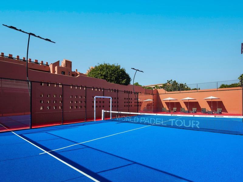 Moderner Tennisplatz mit blauem Belag und roten Wänden, umgeben von Zäunen und Kläranlagen.