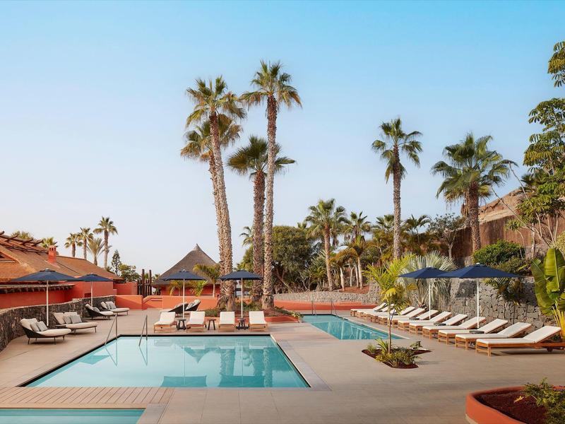 Außerhalb des Hotels mit glattem Wasserpool, Sonnenliegen und hohen Palmen bei klarem Himmel.