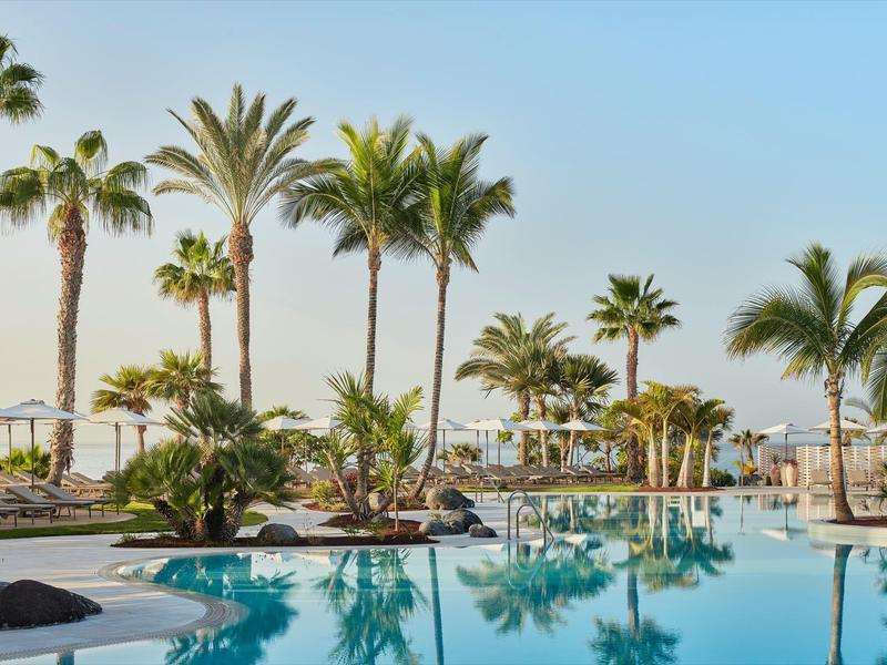 Großer Außenpool mit Palmen, Sonnenliegen und klarem blauem Himmel im Hintergrund.