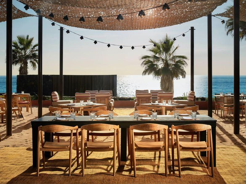 Strandrestaurant mit Holztischen, Stühlen, Palmen und Meer im Hintergrund bei Sonnenuntergang.