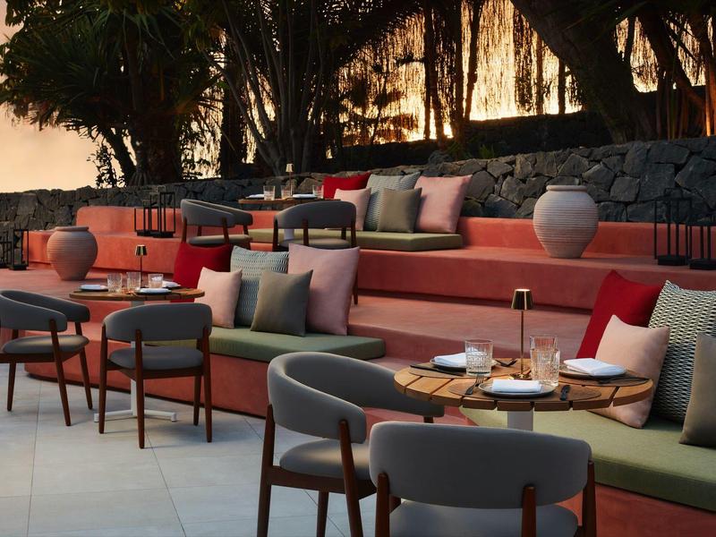 Gemütliche Außenlounge mit roten und grauen Polstern, Tischen und Dekoration in warmem Licht.