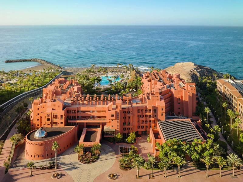Großes rotes Gebäude am Meer mit Palmengarten, vor blauem Wasser und klarem Himmel.