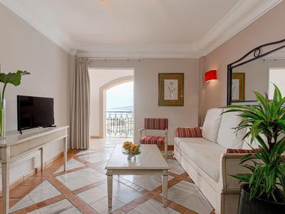 Lichte woonkamer met bank, fauteuils, tv, planten en balkon met zeezicht.