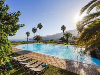 Piscine extérieure avec chaises longues, palmiers et vue sur la mer au coucher du soleil.