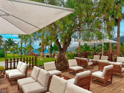 Terrasse mit gepolsterten Stühlen und Tischen unter Bäumen mit Blick auf den Pool