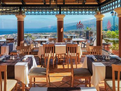 Terrasse de restaurant avec tables et chaises en bois, vue sur la mer et la ville sous un ciel bleu.