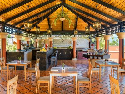 Ruime openluchtbar met houten plafond, tafels, stoelen en bar in een tropische omgeving.