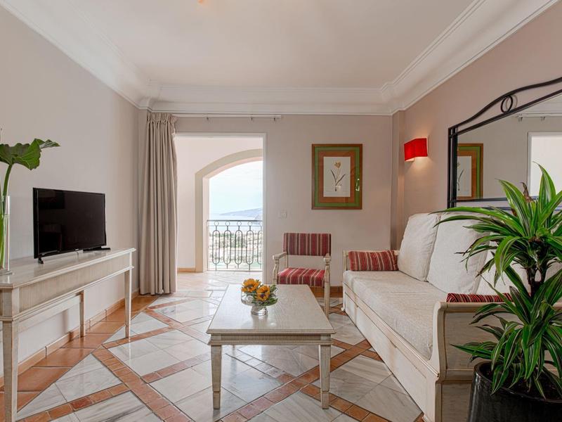 Lichte woonkamer met bank, fauteuils, tv, planten en balkon met zeezicht.