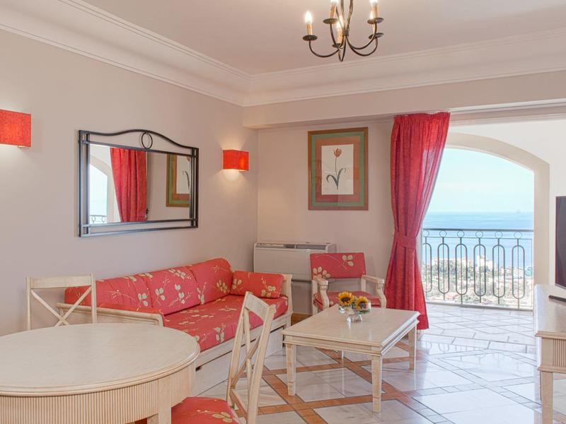 Lichte woonkamer met rode bank, witte tafel en zeezicht vanaf balkon.