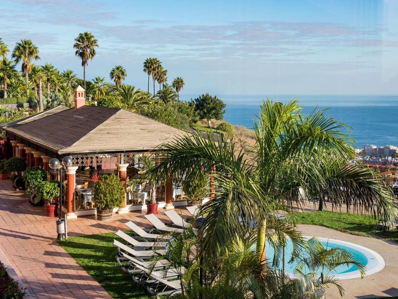 Hotel met zwembad, palmbomen en uitzicht op zee onder een helderblauwe lucht.