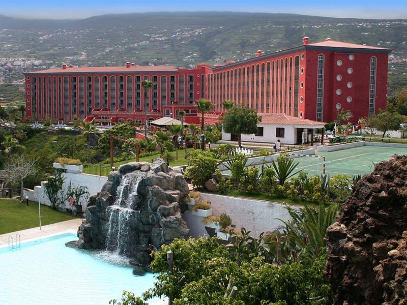 Groot rood hotel met tennisbanen en watervalzwembad in een groen, heuvelachtig gebied.