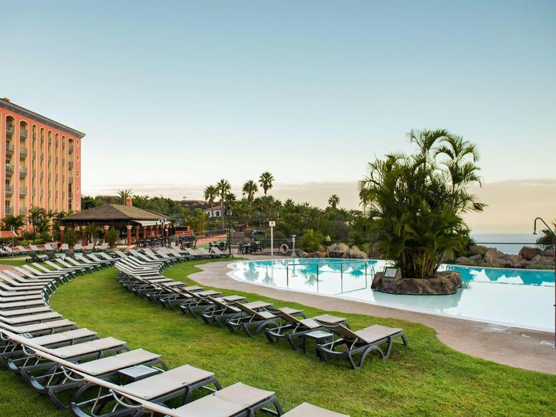 Hoteltuin met zwembad, veel ligstoelen en palmbomen onder een heldere hemel.