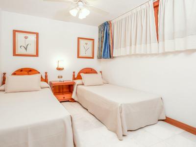 Einfaches Schlafzimmer mit zwei Einzelbetten, Holzmöbeln, weißen Wänden und Vorhängen.