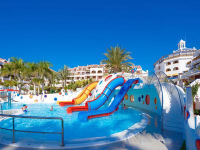 Bunter Wasserrutschenbereich mit Pool, umgeben von Hotelgebäuden und Palmen unter klarem blauem Himmel.