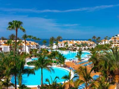 Große blaue Pools umgeben von Palmen, weißen Gebäuden und blauem Himmel in sonnigem Resort.