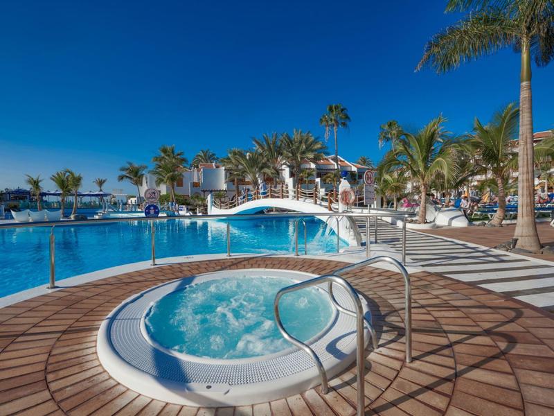 Freiluft-Whirlpool vor großem Pool, umgeben von Palmen und weißen Gebäuden unter blauem Himmel.