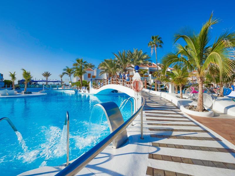 Großer Außenpool mit blauer Brücke und Palmen unter klarem, blauem Himmel.