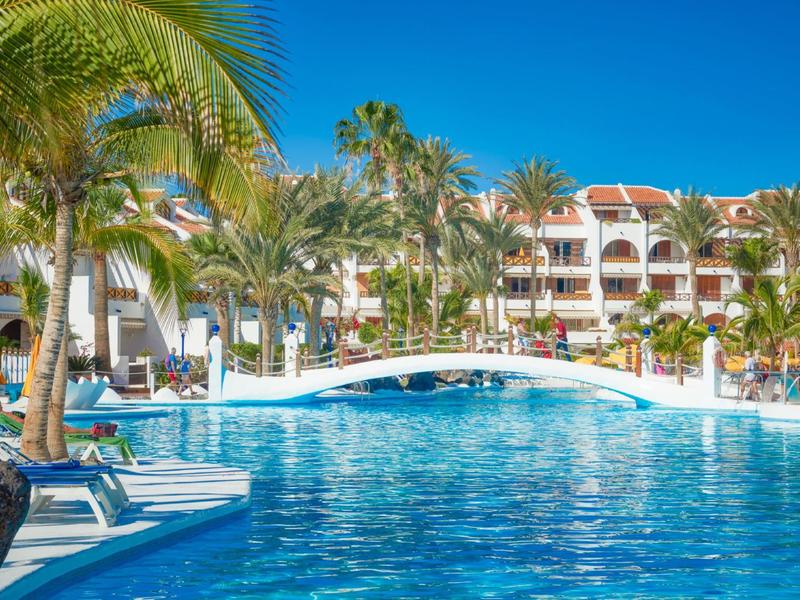 Großer blauer Pool mit weißer Brücke, Palmen und mehrstöckigen Gebäuden im Hintergrund bei klarem Himmel.