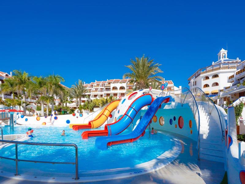 Bunter Wasserrutschenbereich mit Pool, umgeben von Hotelgebäuden und Palmen unter klarem blauem Himmel.