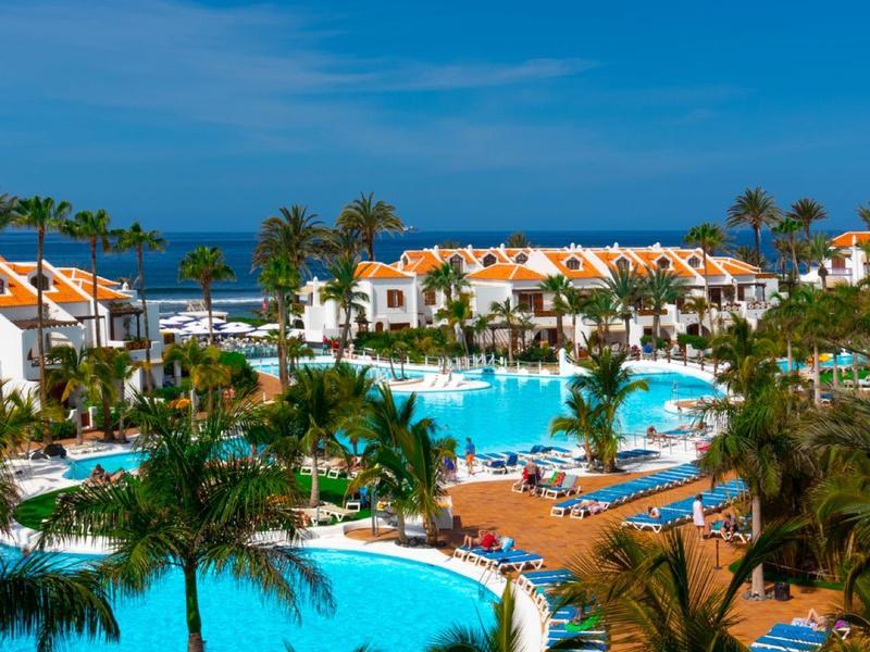 Große Poollandschaft mit Palmen, weiß-orangenen Häusern und blauem Himmel am Meer.