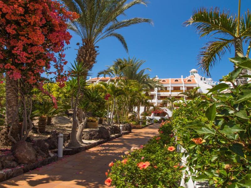 Weg mit Palmen und bunten Blumen vor einem weißen Hotelgebäude unter klarem blauem Himmel.