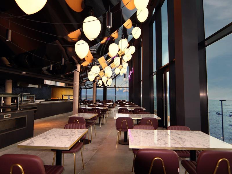 Modern restaurant with large windows and decorative lights overlooking the sea.