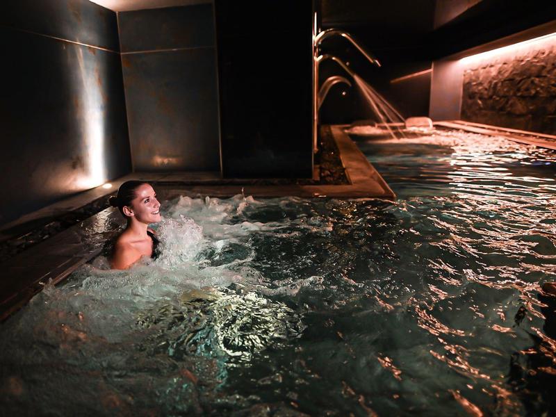 Person relaxing in a lit indoor pool with waterfalls in a hotel setting.