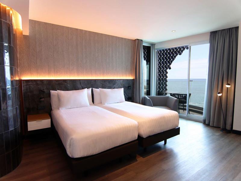 Modern hotel room with two beds, seating area and sea view through large glass windows.