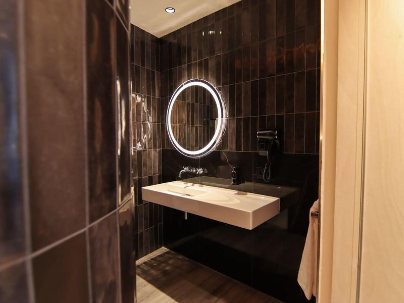 Modern hotel bathroom with illuminated oval mirror and white sink against dark wall.